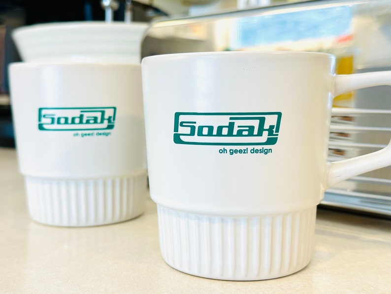May include: Two white ceramic mugs with a green logo that says "Sodak oh geez! design". One mug has a handle and the other does not.