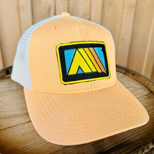 May include: A peach-colored trucker hat with a white mesh back. The hat has a black and yellow patch with a blue, yellow, and orange graphic of a mountain range.