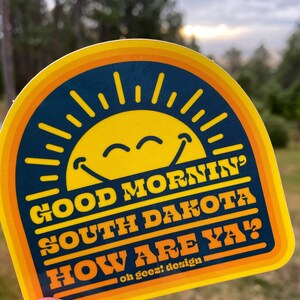 May include: Yellow and blue sticker with a smiling sun and the text "Good Morning South Dakota How Are Ya?" with the words "oh geez! design" at the bottom.