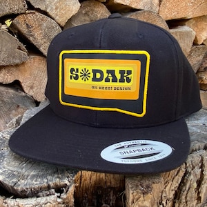 May include: Black baseball cap with a yellow and orange patch that says "S*DAK OH GEEZ! DESIGN" and a white label on the back that says "THE AUTHENTIC SNAPBACK YUPOONG SINCE 1974"