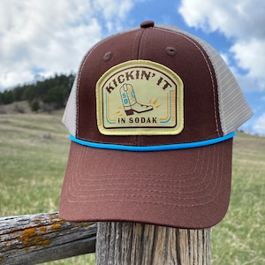 May include: Brown and beige trucker hat with a blue trim. The hat has a patch on the front that says "Kickin' It In  Sodak" with a cartoon cowboy boot.