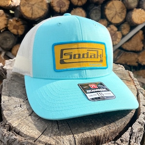 May include: A light blue and white trucker hat with a yellow and blue patch that says "Sodak!" on it. The hat has a Richardson label on the side.