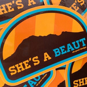 May include: A sticker featuring a mountain silhouette and the text "She's a beaut" in blue lettering. The sticker has a brown background with orange and yellow stripes and a blue border.