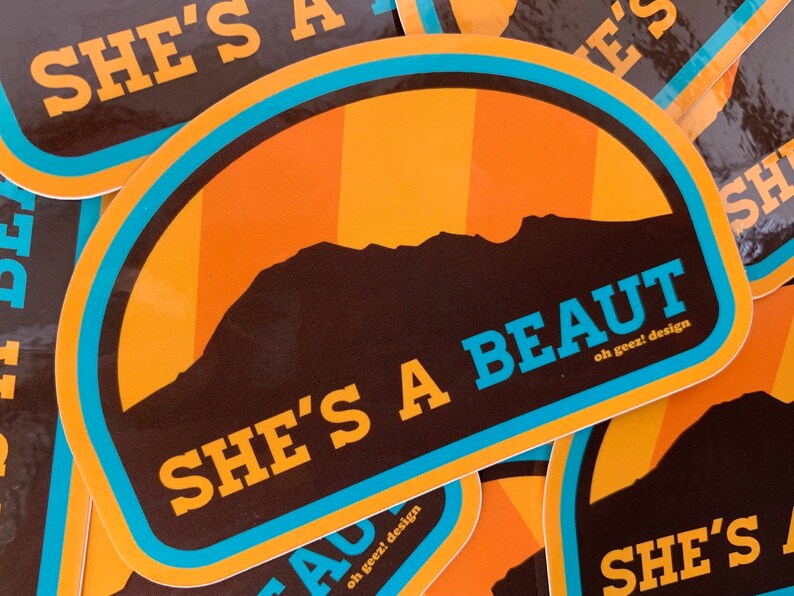 May include: A sticker featuring a mountain silhouette and the text "She's a beaut" in blue lettering. The sticker has a brown background with orange and yellow stripes and a blue border.