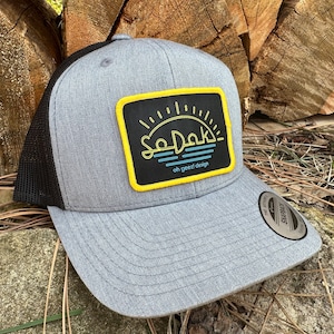 May include: A gray and black trucker hat with a yellow and black patch that says "SoDak!" with a sunset and waves design. The patch also says "oh geet! design".