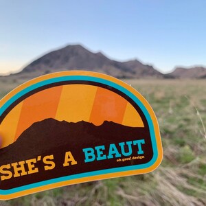 May include: A yellow and blue sticker with a brown silhouette of a mountain range and the text "SHE'S A BEAUT oh geez! design".