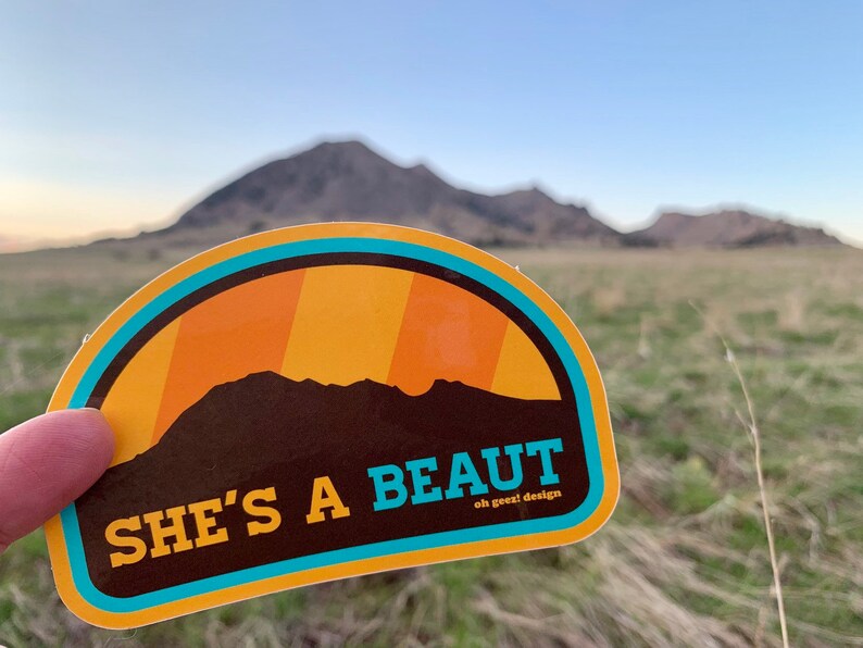 May include: A yellow and blue sticker with a brown silhouette of a mountain range and the text "SHE'S A BEAUT oh geez! design".