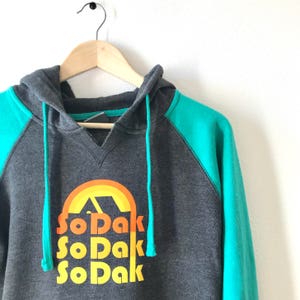 May include: A gray and teal hooded sweatshirt with a graphic of a sunset and the text "So Dak" repeated three times.