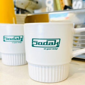 May include: White ceramic mug with a green logo that says "Sodak! oh geezi design".