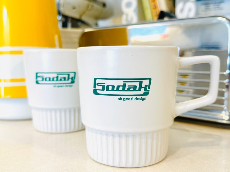 May include: White ceramic mug with a green logo that says "Sodak! oh geezi design".
