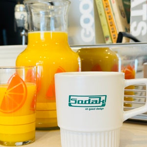 May include: A white ceramic mug with a green logo that says "Sodak oh geez! design". The mug is sitting on a counter next to a jug and glass of orange juice.