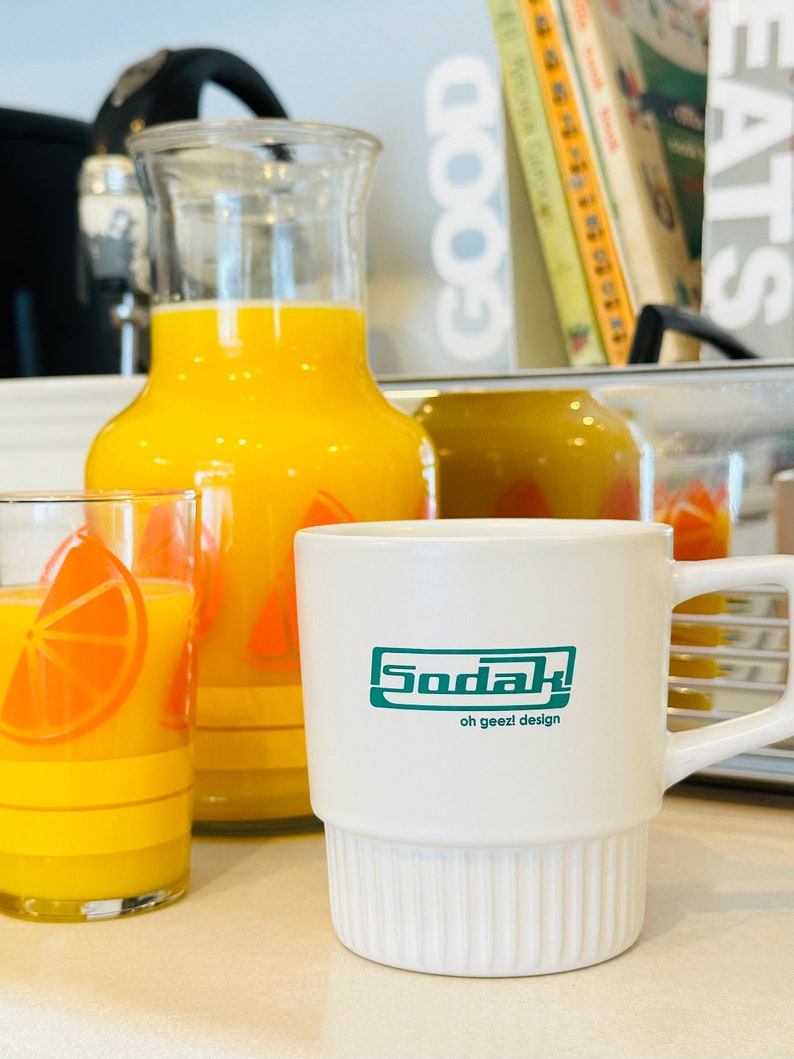 May include: A white ceramic mug with a green logo that says "Sodak oh geez! design". The mug is sitting on a counter next to a jug and glass of orange juice.