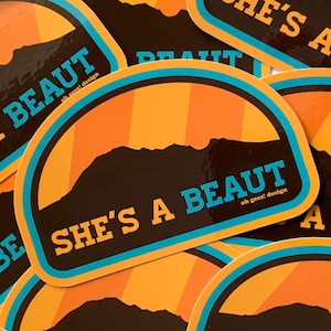May include: A sticker featuring a mountain silhouette and the text "She's a beaut oh geez! design" in a retro style with orange, yellow, blue, and black colours.