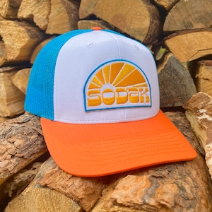 May include: A white and blue trucker hat with an orange bill. The hat has a yellow embroidered patch with a sun and the word "SODAK" on it.