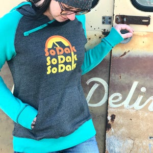 May include: A gray hooded sweatshirt with teal blue sleeves and a teal blue bottom band. The sweatshirt has a graphic print of a yellow sun with a mountain inside and the text "SoDak SoDak SoDak" in yellow.