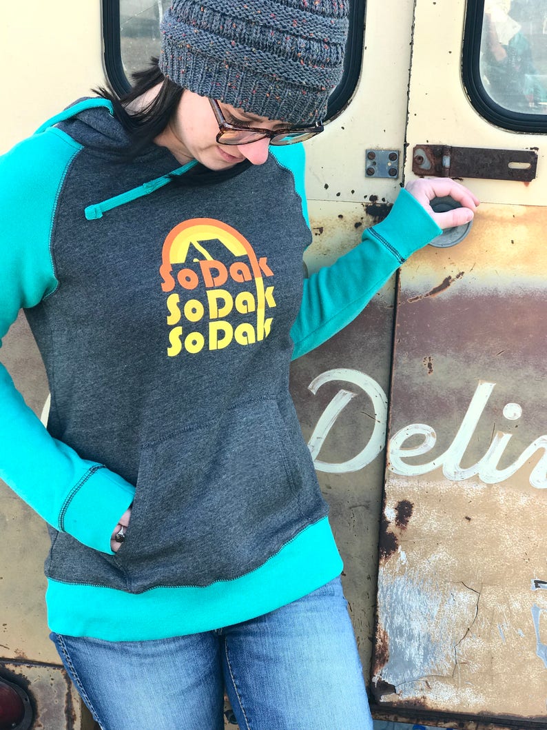 May include: A gray hooded sweatshirt with teal blue sleeves and a teal blue bottom band. The sweatshirt has a graphic print of a yellow sun with a mountain inside and the text "SoDak SoDak SoDak" in yellow.
