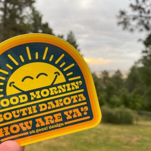 May include: A yellow and orange sticker with a smiling sun and the text "Good Morning South Dakota How Are Ya?"  oh geez! design