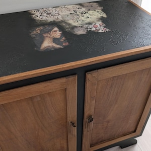 Puede incluir: Un mueble de madera con dos puertas que presenta paneles de veta natural y un marco negro. La superficie superior está pintada de negro con un patrón en relieve y una imagen decorativa transferida de un perfil femenino con motivos florales.