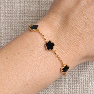 Op de afbeelding: Een gouden schakelarmband met drie gelijkmatig verdeelde bloemvormige bedels. Elke bedel heeft een gladde, effen zwarte inleg in een gepolijste goudkleurige metalen rand. De fijne schakelketting verbindt elk bloemmotief in een herhalend patroon.
