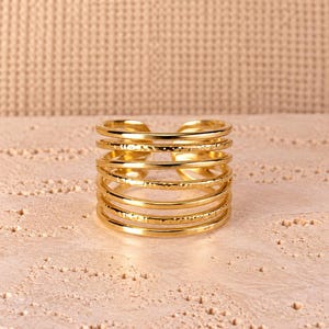 Könnte beinhalten: Ein goldfarbener Ring mit einem mehrreihigen Design aus sechs parallelen, dünnen, kreisförmigen Drähten. Die Bänder wechseln zwischen glatten, polierten Oberflächen und strukturierten, gehämmerten Oberflächen.
