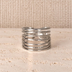 Op de afbeelding: Een brede zilverkleurige ring met een ontwerp van meerdere banden, bestaande uit verschillende dunne, gestapelde metalen draden. Het oppervlak van de banden heeft een subtiele gehamerde textuur, wat zorgt voor een reflecterende afwerking.