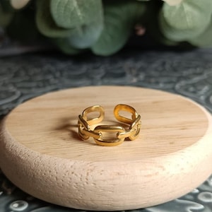 Può includere: Un anello a catena dorata su una superficie di legno. L'anello è realizzato con un design a catena spessa e robusta.