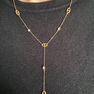 Op de afbeelding: Een goudkleurige ketting met een Y-vormige hanger. De ketting heeft ovale schakels en kleine, heldere edelstenen. De hanger heeft een bungelende ketting met een enkele edelsteen en een ovale bedel aan het einde.