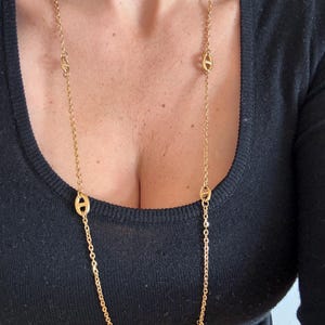 Peut inclure: Un long collier doré avec une chaîne délicate et plusieurs maillons ovales. Le collier est porté sur un haut noir. Les maillons sont espacés uniformément le long de la chaîne, créant un design simple mais élégant.