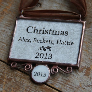 Puede incluir: Adorno navide&ntilde;o rectangular con una cinta marr&oacute;n. El adorno tiene la palabra "Christmas" y los nombres "Alex, Beckett, Hattie", junto con el a&ntilde;o "2013". Tiene un borde decorativo y una peque&ntilde;a etiqueta circular con "2013".
