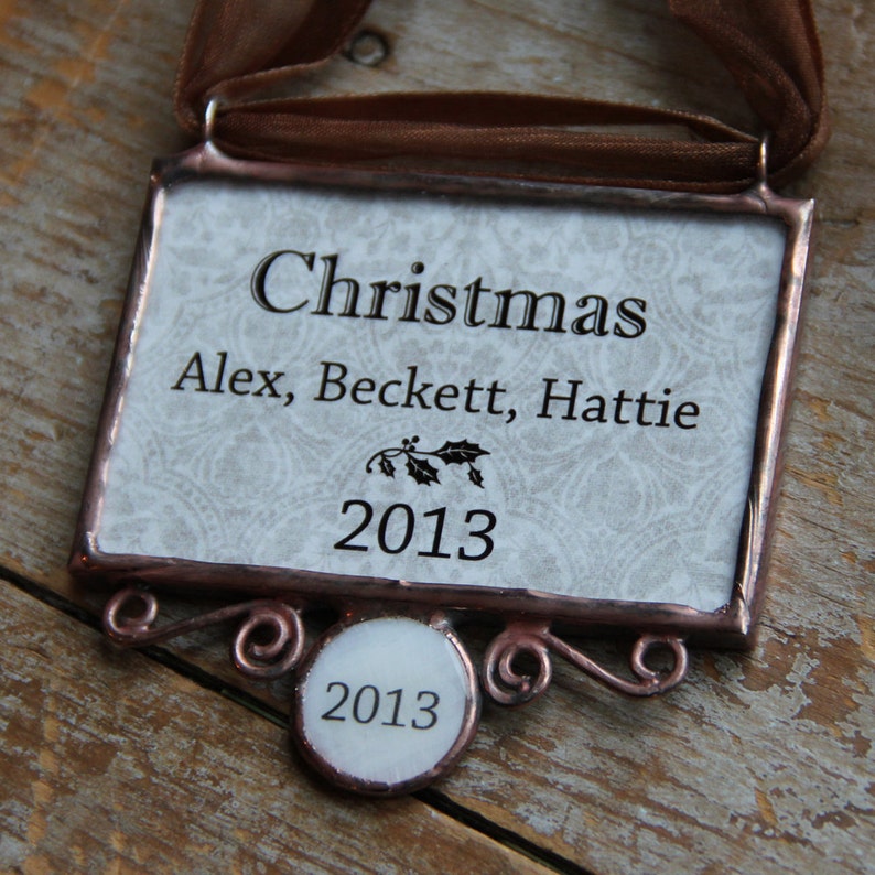 Puede incluir: Adorno navide&ntilde;o rectangular con una cinta marr&oacute;n. El adorno tiene la palabra "Christmas" y los nombres "Alex, Beckett, Hattie", junto con el a&ntilde;o "2013". Tiene un borde decorativo y una peque&ntilde;a etiqueta circular con "2013".