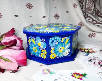 May include: A blue wooden octagonal box with a floral design painted on it. The flowers are yellow, blue, and white. The box is sitting on a white surface with a few pink roses in the background.
