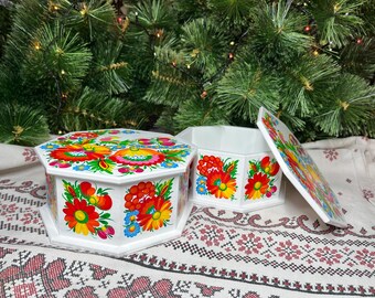 May include: White, hexagonal decorative boxes with lids, adorned with vibrant floral patterns in red, yellow, blue, and green. The boxes are displayed on a patterned cloth, with a Christmas tree in the background.