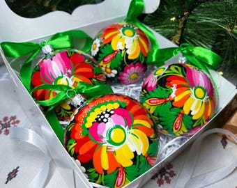 May include: Five hand-painted Christmas baubles in a white box. Each glass bauble features a vibrant floral design in red, pink, yellow, and green, with a black background. Green ribbons are tied to the top.