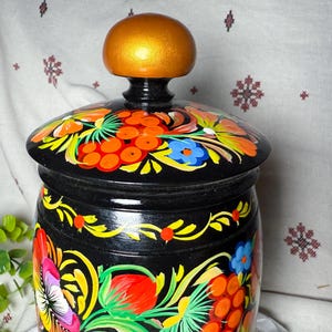May include: A black, hand-painted wooden jar with a lid. The jar is adorned with vibrant floral and berry designs in red, orange, yellow, and blue. The lid features a gold-colored knob. This decorative jar is a unique piece of folk art.