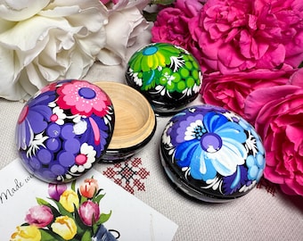 May include: Hand-painted wooden trinket boxes with floral designs in purple, pink, blue, and green. The boxes are black with a wooden interior. A card with a floral bouquet and the words "Made with" is in the foreground.