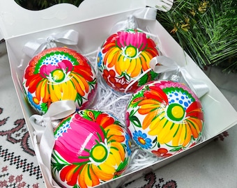 May include: Four hand-painted Christmas baubles in a white box. The baubles are round and feature a white background with colourful floral designs. The baubles are decorated with pink, yellow, red, green, and blue paint.