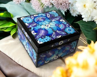 May include: A black wooden box with a glossy finish, decorated with a vibrant floral design in shades of blue and purple. The flowers are painted on the lid and sides, creating a decorative storage item.