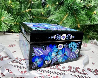 May include: A black wooden box with a hinged lid, painted with a floral design in shades of blue and purple. The box is decorated with white flowers and leaves.