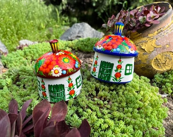 May include: Two hand-painted, decorative trinket boxes shaped like small houses. The lids are adorned with red berry and floral patterns. The houses have white walls, green doors, and small windows, set against a backdrop of green foliage.