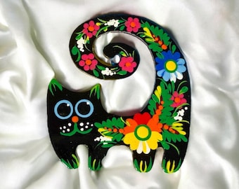 May include: A whimsical, black wooden cat figurine with a curled tail, adorned with colorful floral patterns. The cat features large blue eyes, a green and white dotted mouth, and green accents on its paws and ears. The flowers include pink, yellow, and blue.