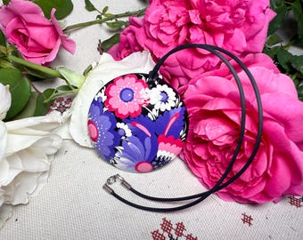 May include: A round pendant necklace with a black cord. The pendant features a floral design in pink, purple, and white on a black background. The necklace is set against a backdrop of pink and white roses.