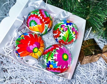 May include: Four round Christmas baubles with a white background and colourful floral designs. The baubles are in a white box with white shredded paper.