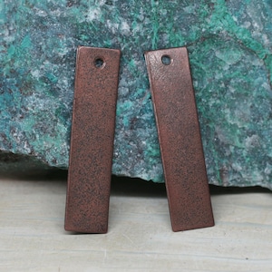 May include: Two rectangular, copper-colored metal pendants with a textured finish. Each pendant has a small hole at the top. The pendants are displayed against a background of green and blue stone.