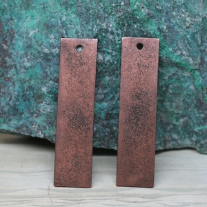 May include: Two rectangular copper metal tags with a hole punched in the top. The tags have a distressed, aged, and rustic finish.