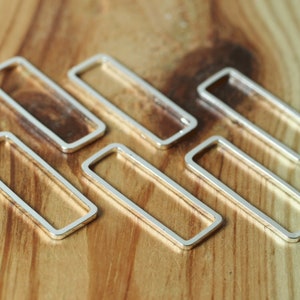 May include: Six silver rectangular jewellery findings are arranged on a wooden surface. Each finding is a hollow rectangle with rounded corners. The findings are made of a shiny, metallic material, and are evenly spaced.