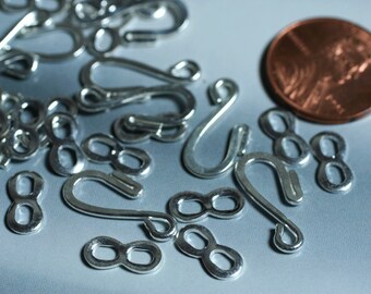 May include: Close-up of silver-colored jewelry findings, including hook clasps and figure-eight connectors. A U.S. penny is included for size reference. The findings are scattered on a light gray surface, showcasing their metallic sheen.