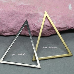 May include: Two open-frame triangle pendants, one gunmetal and one raw brass, are displayed against a pink and gray background. The gunmetal triangle is a dark gray color, while the brass triangle has a golden hue. Text on the image reads "gun metal" and "raw brass."