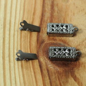 May include: Close-up of jewelry clasps and clips. The clasps are rectangular with a decorative openwork design. The clips are small and have a simple, functional design. All pieces are silver-toned and sit on a wooden surface.