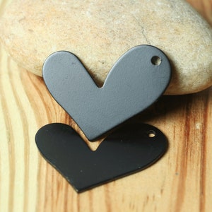 May include: Two heart-shaped metal pendants, one gray and one black, each with a small hole at the top. The pendants are on a wooden surface with a stone in the background. The hearts are likely for jewelry making or crafts.
