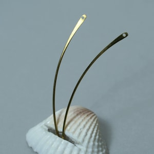 May include: Two slender, curved, gold-colored metal earring components with small holes at the ends. The earrings are displayed on a white seashell against a soft gray background. The design is minimalist and elegant.